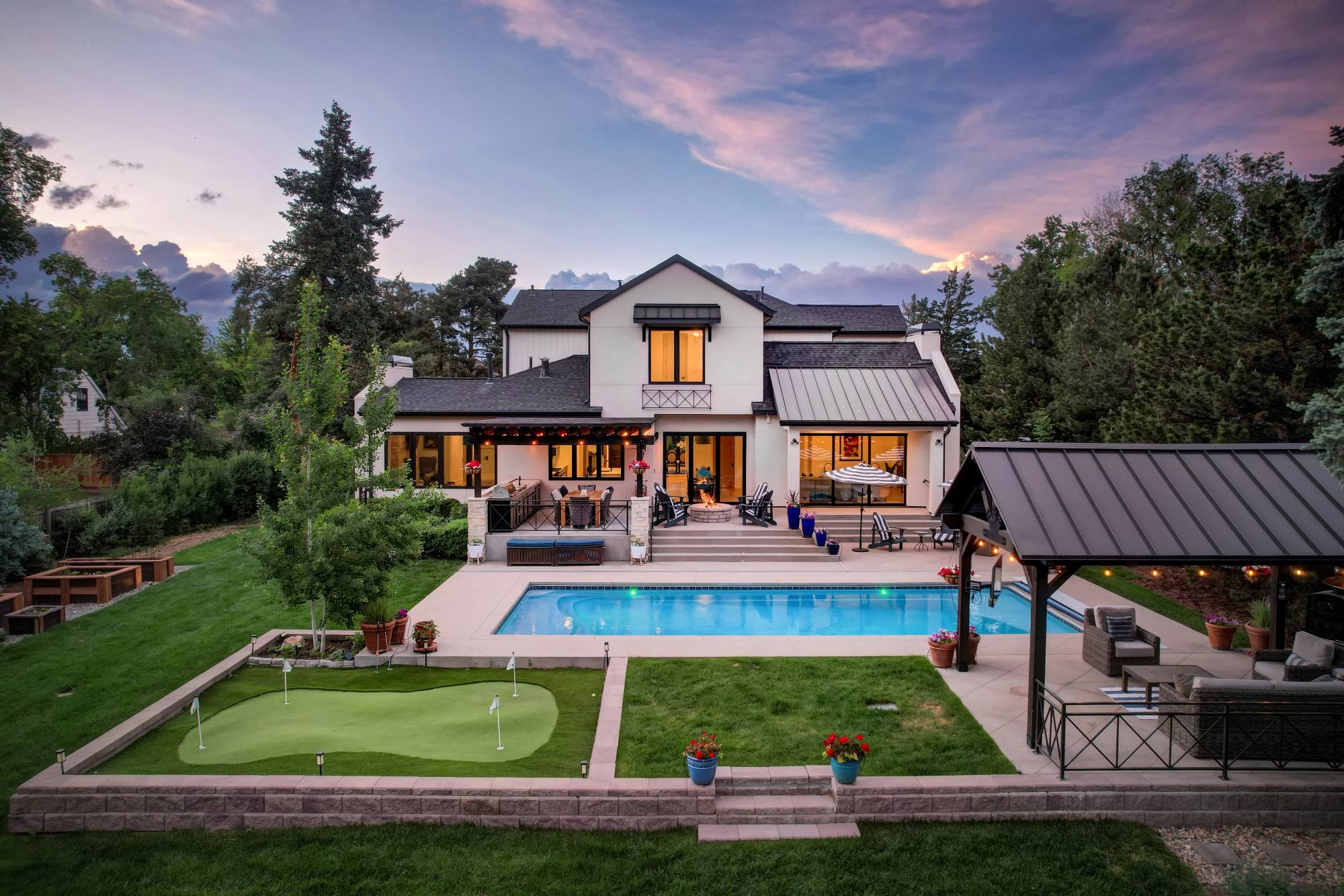 Backyard at twilight with a lit pool, putting green, and a string-lit pergola under a pink sunset